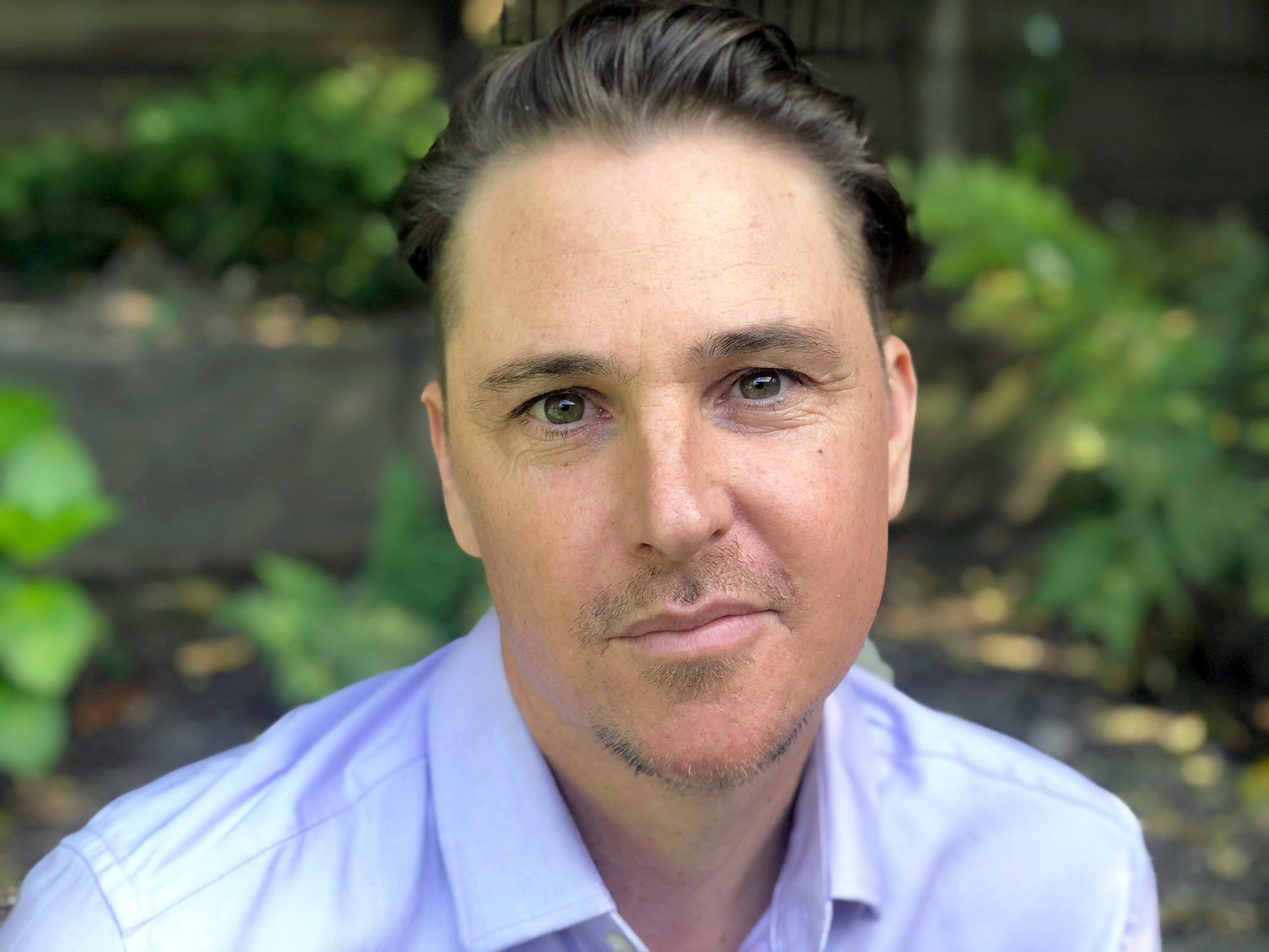 Picutre of a white man with dark brown hair and a goatee. His shirt is a light blue collared shirt, and he's outside in front of greenery.